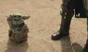 Grogu (Baby Yoda) in a tan robe stands on a sandy desert beside a person wearing dark Mandalorian armor. He raises a hand as if waving.