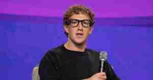 Male speaker with curly hair and glasses sits on stage holding a microphone during a talk against a purple backdrop.