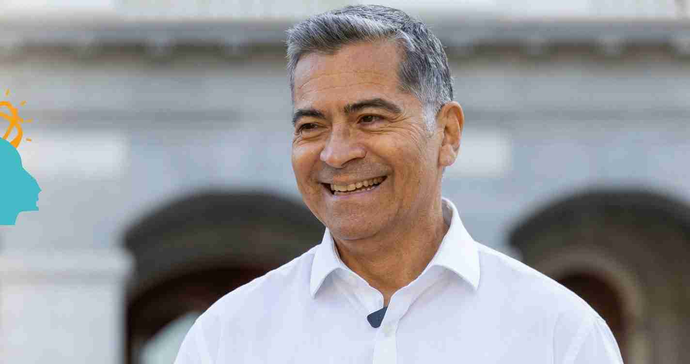 Smiling middle-aged man with gray hair in a white shirt, outdoors, with a turquoise logo partially visible on the left.