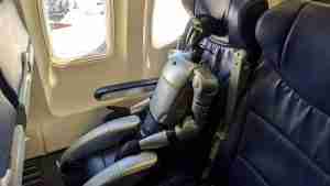 Small humanoid robot seated in an airplane seat by a window, as if a passenger, with blue leather seats nearby.