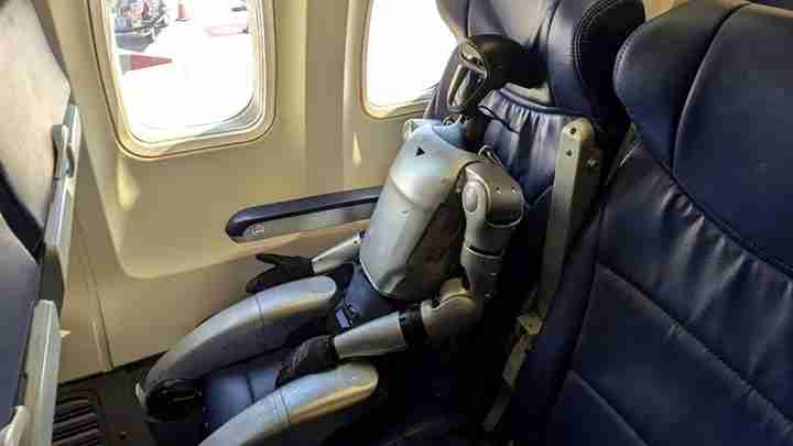 Small humanoid robot seated in an airplane seat by a window, as if a passenger, with blue leather seats nearby.