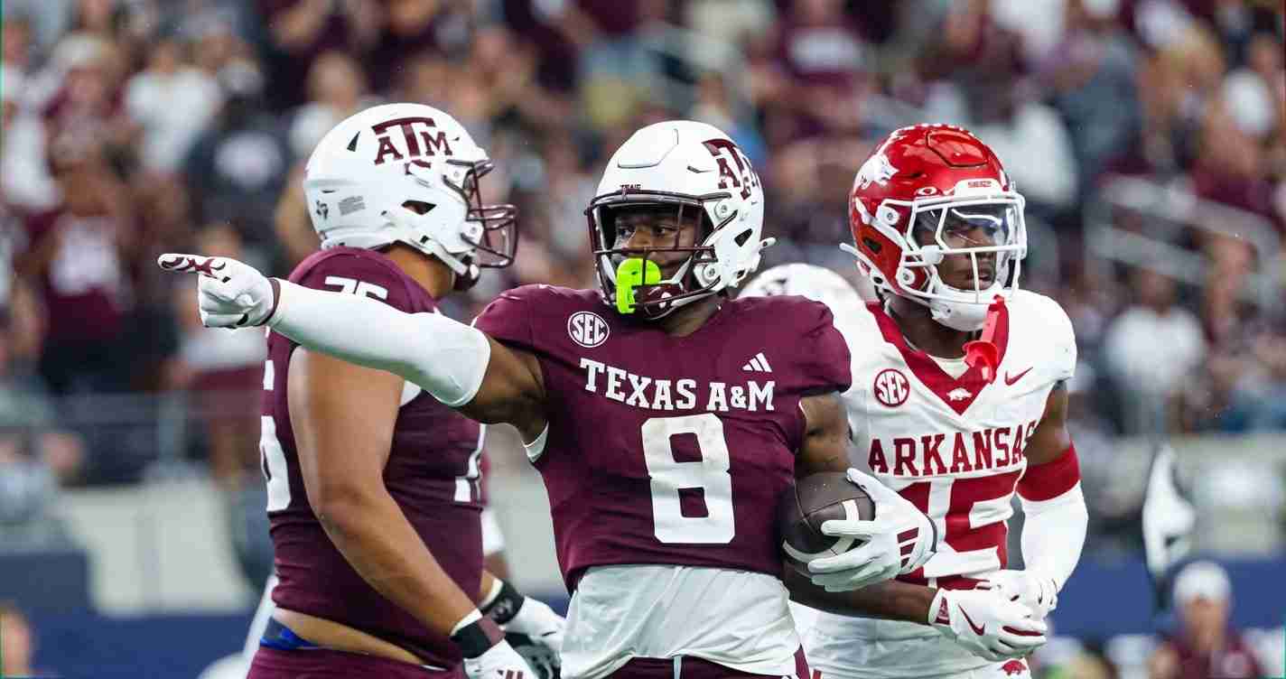 Texas A&M running back in maroon jersey with ball, pointing downfield as an Arkansas defender closes in
