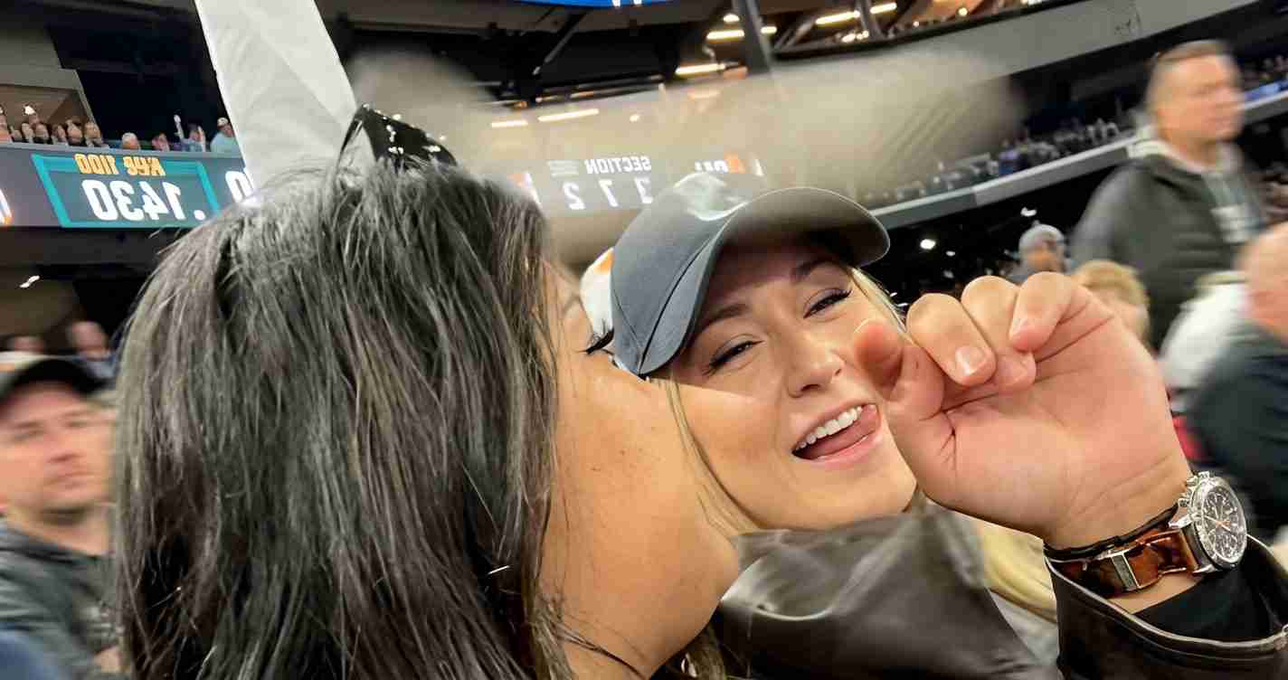 Two friends in a stadium share a kiss on the cheek, with one wearing a gray cap and a wristwatch visible on her wrist.