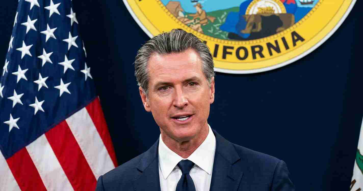 Male official in a dark suit speaks at a press conference, with the US flag and California state seal behind him.