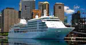 A large white cruise ship docked at a marina with a city skyline of tall buildings in the background.