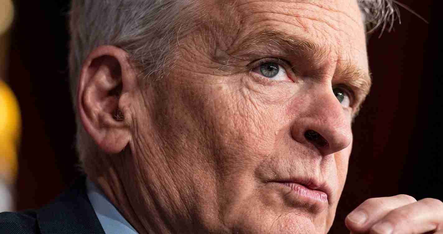 Close-up of a man with gray hair and blue eyes, resting his chin on his hand in thought.