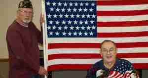 Two older men pose with a large American flag, one standing in a maroon sweater and cap, the other seated in a patriotic shirt.