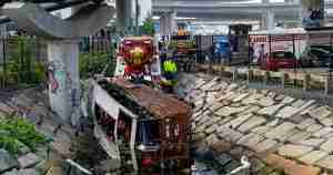 Bus crash in a rocky drainage canal under a highway, with fire trucks and responders on scene to assist occupants and secure the area.
