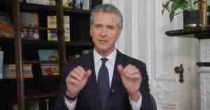 Man in a dark suit and blue tie speaks, hands raised in a gesture, seated in a room with white paneling and a bookshelf in the background