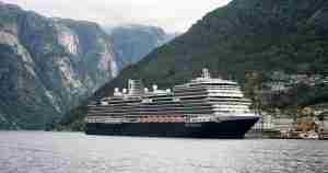 Large cruise ship in a fjord, with steep forested mountains and a small shoreline village nearby.
