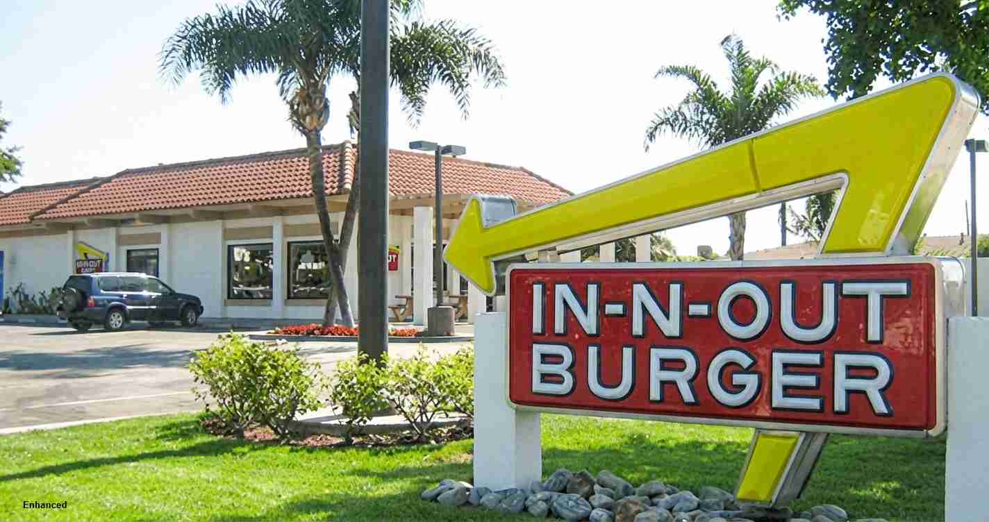 Bright In-N-Out Burger sign with a large yellow arrow in the foreground, a fast-food building and palm trees behind on a sunny day.