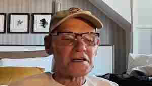 Older man wearing a tan cap and glasses speaks to the camera in a hotel room with framed dog prints in the background.