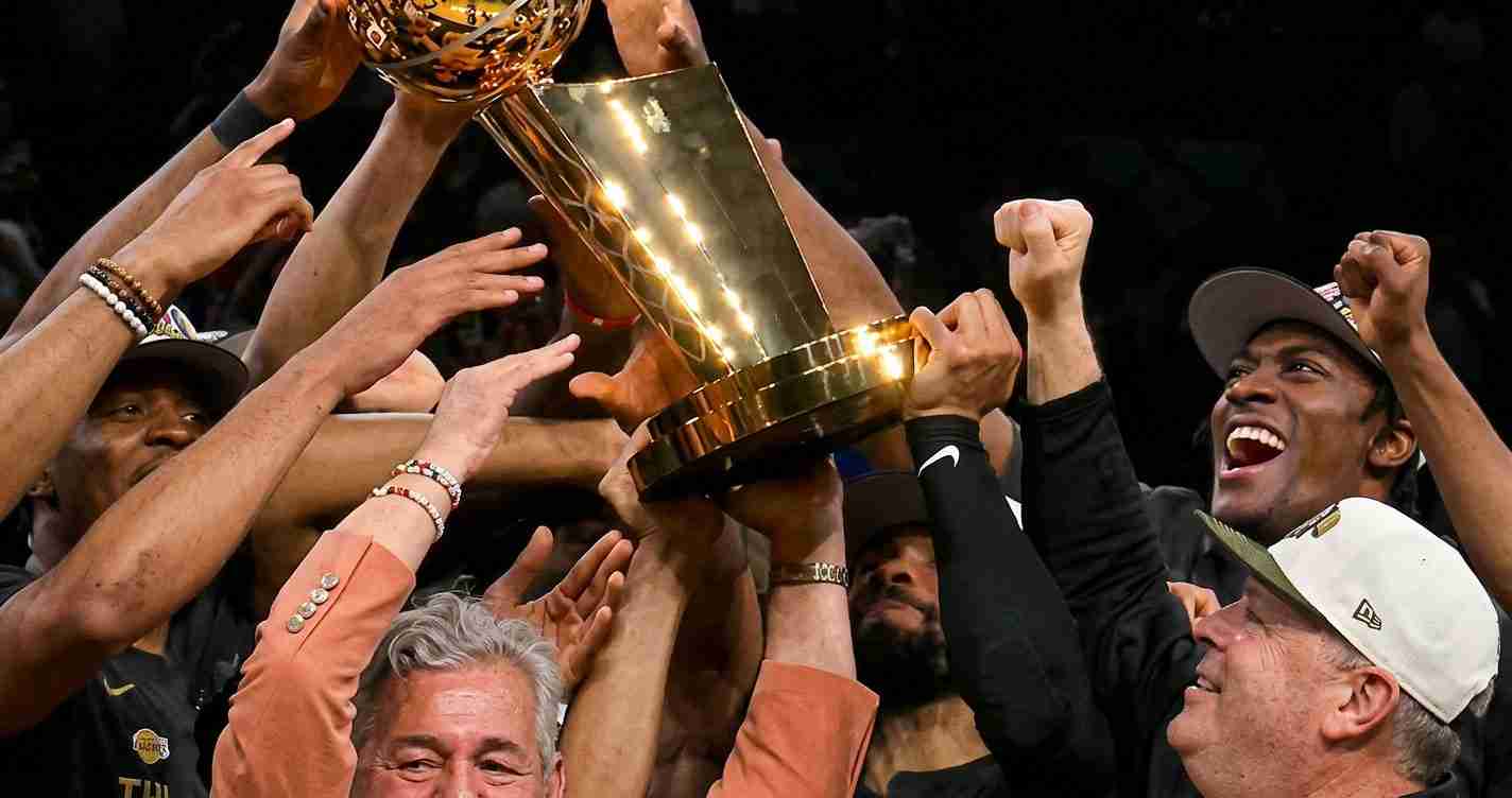 Athletes raise a large golden trophy in celebration, their hands reaching up together.