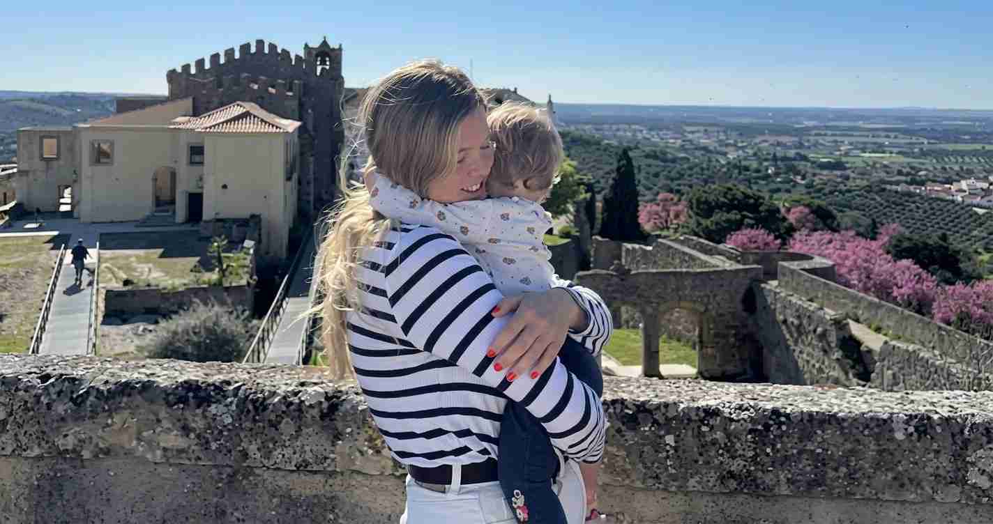 Woman in a striped shirt hugs a child against a stone fortress wall with a scenic valley beyond, sunny day.