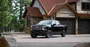 Black pickup truck parked in a driveway beside a rustic stone-and-wood house in a wooded setting.