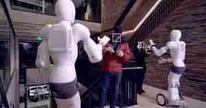 Two white humanoid robots high-five a man in a red jacket in a modern showroom or lobby setting, with a wooden wall behind them.