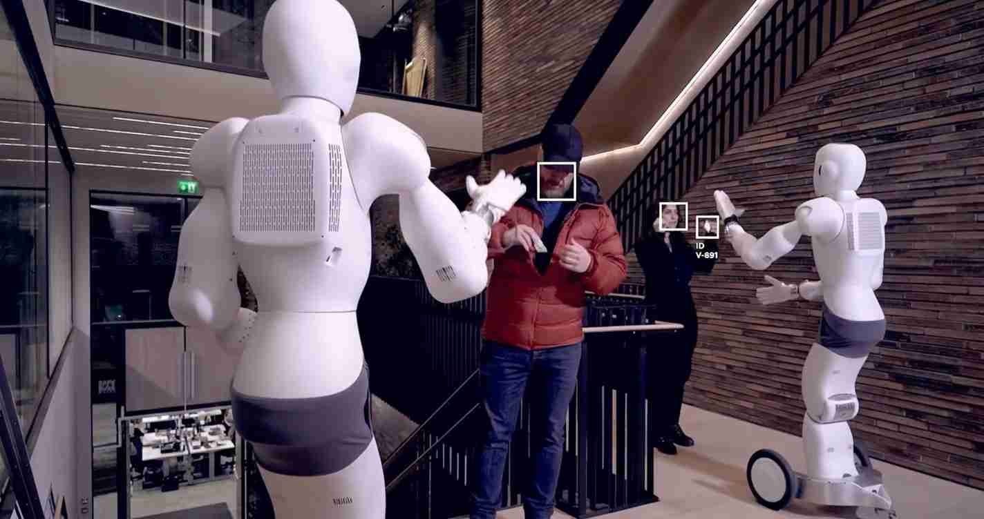 Two white humanoid robots high-five a man in a red jacket in a modern showroom or lobby setting, with a wooden wall behind them.