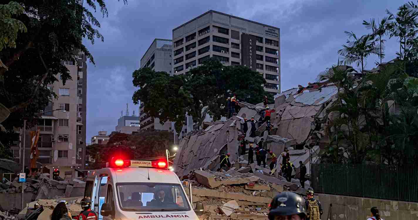 Emergency responders climb a collapsed concrete building amid rubble, with an ambulance and flashing red lights in the foreground.