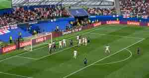 Soccer match near the goal with players in white defending and dark kits attacking; crowd in stands behind the goal.