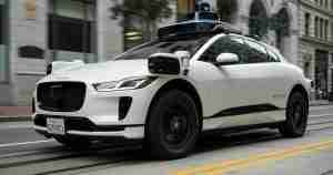 White Jaguar SUV equipped with Waymo self-driving sensors on the roof driving through a city street.