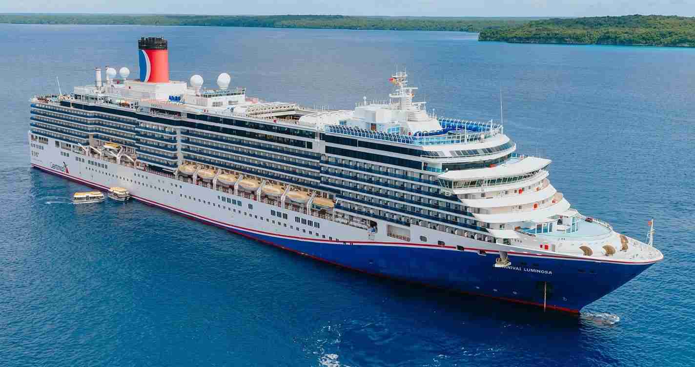 A large white and blue cruise ship with many decks and a red funnel, sailing on calm blue sea with a distant green coastline.