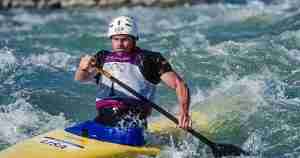 Male kayaker in USA gear paddles through white-water in a yellow kayak, helmet on, brace on a rough river.