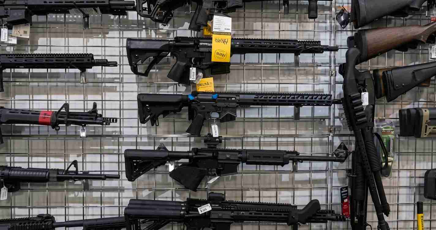 Assorted rifles and shotguns displayed on a metal grid wall in a gun shop with price tags visible.