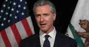 Unidentified man in a dark suit and tie speaks with the US and California flags in the background.