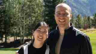 Two adults standing close together outdoors in a park, smiling at the camera, with trees and mountains in the background.