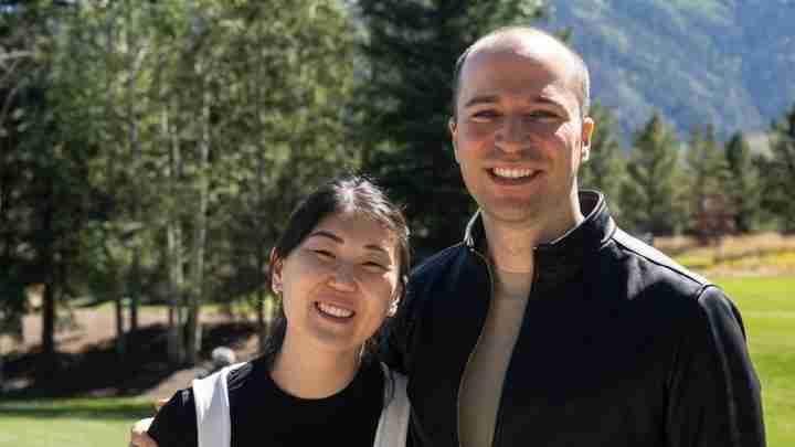 Two adults standing close together outdoors in a park, smiling at the camera, with trees and mountains in the background.