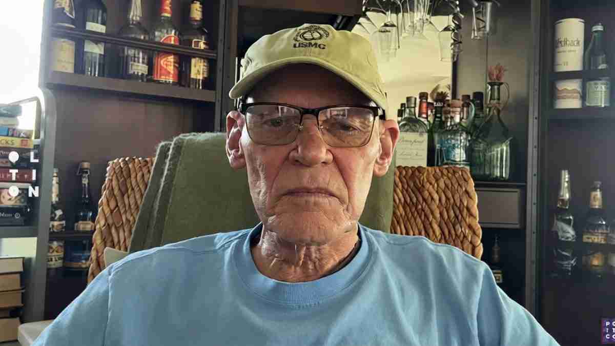 Older man wearing a USMC cap and glasses sits in a cozy bar or lounge with bottles on shelves in the background.