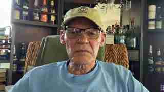Older man wearing a USMC cap and glasses sits in a cozy bar or lounge with bottles on shelves in the background.