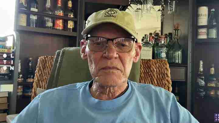 Older man wearing a USMC cap and glasses sits in a cozy bar or lounge with bottles on shelves in the background.