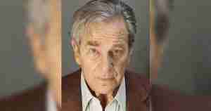 Portrait of an older man with gray hair and a serious expression, wearing a brown blazer over a white collared shirt.
