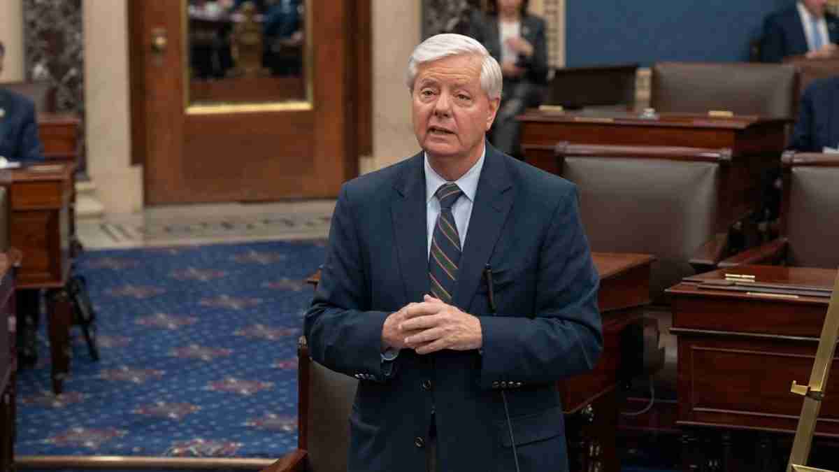 Older man in a navy suit and striped tie stands in a legislative chamber, speaking to others nearby.