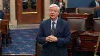Older man in a navy suit and striped tie stands in a legislative chamber, speaking to others nearby.