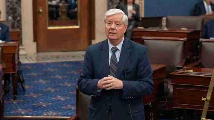 Older man in a navy suit and striped tie stands in a legislative chamber, speaking to others nearby.