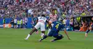 Soccer player in white-and-red kit challenges for the ball as a blue-jersey defender slides in during a crowded stadium match.