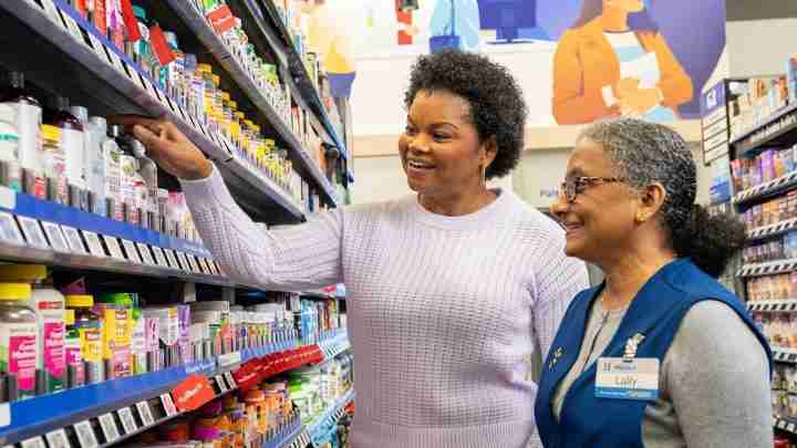 Two women in a pharmacy aisle; a shopper reaches for a product while a staff member in a blue vest watches and smiles.