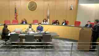 Officials wearing masks sit at a curved conference table in a government chamber, California seal and flags visible behind them.